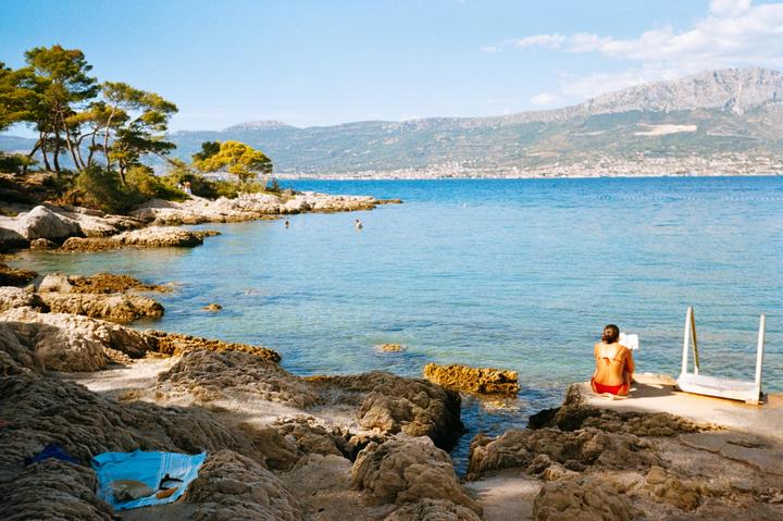 A woman sits on a concrete slab by the sea, with mountains in the distance.