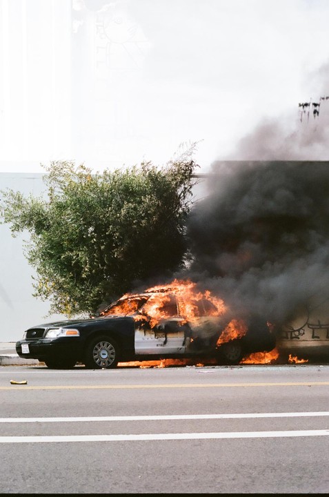 A police car is engulfed in flames and smoke on a street next to a bush.
