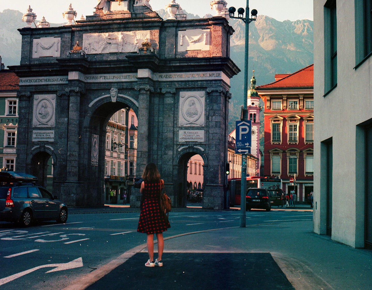 A woman stands in a street looking at a large stone archway with mountains in the background.