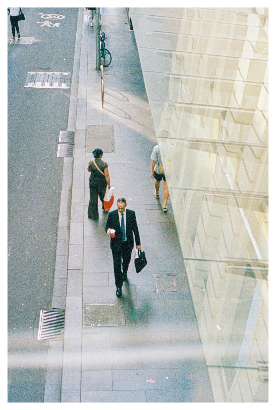 A man in a suit holding a cup and a bag walks on a city sidewalk next to a glass building.