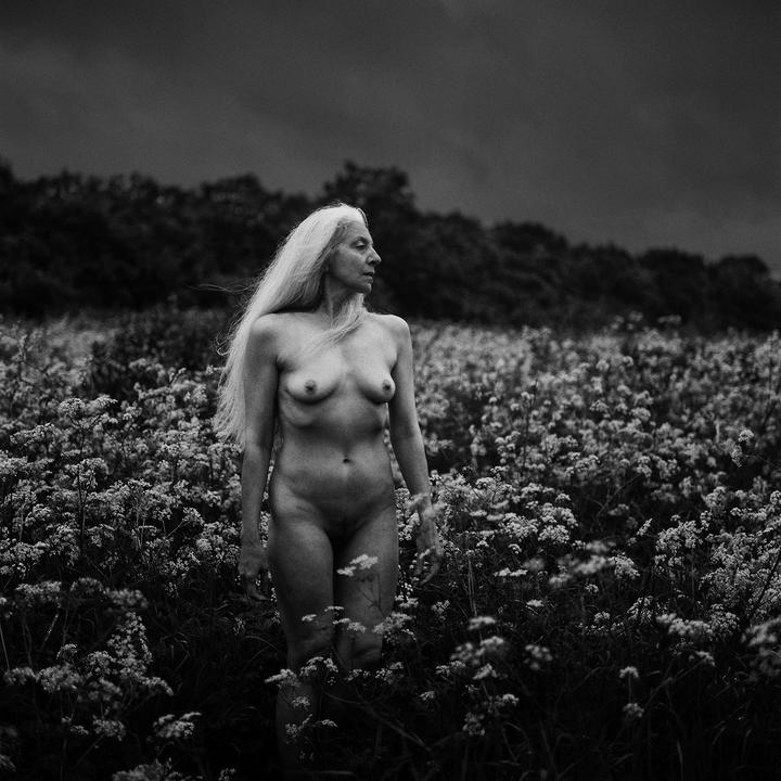 A nude woman with long hair stands in a field of wildflowers under a dark sky.