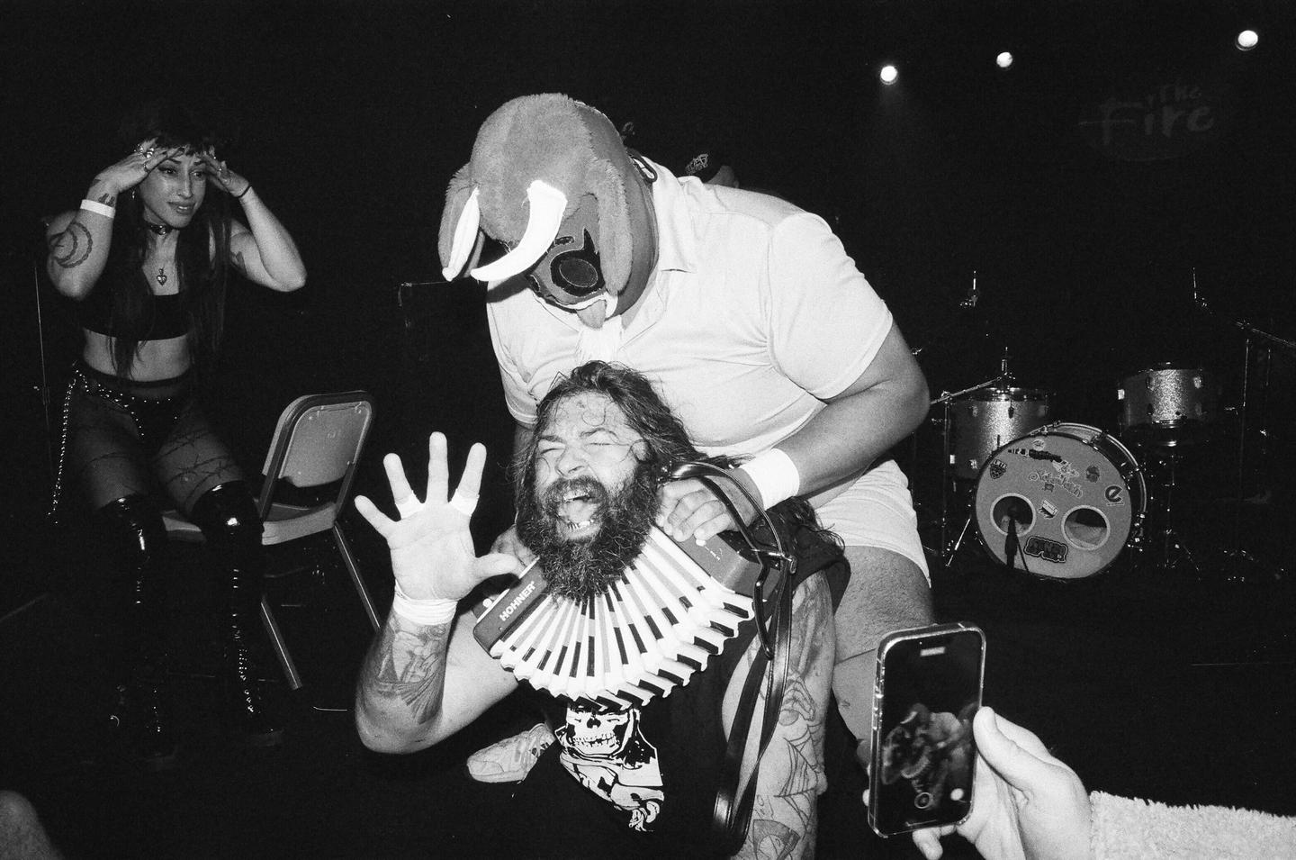 A man in an elephant mask holds another man with a beard and an accordion, while a woman watches and someone records with a phone.