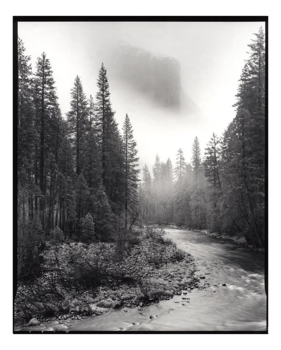 A river flows through a forest with a large rock formation in the background shrouded in fog.