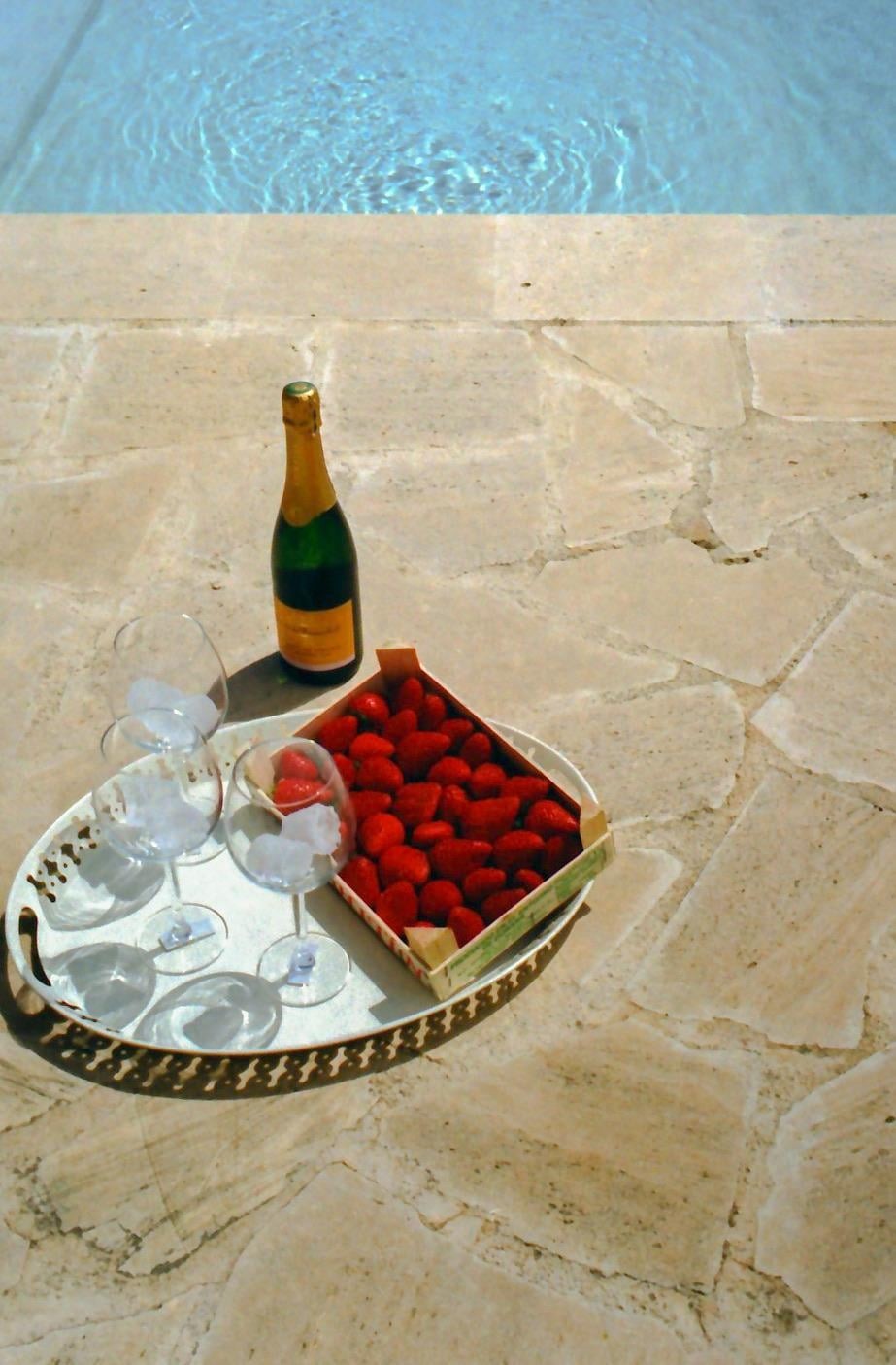 A bottle of champagne, a tray of strawberries, and four glasses with ice sit on a stone patio next to a swimming pool.