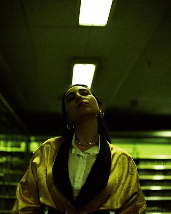 A woman with closed eyes and a gold robe stands under fluorescent lights.