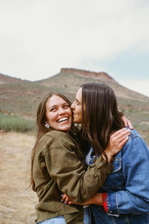 Two women embrace and laugh outdoors with a mountain in the background.