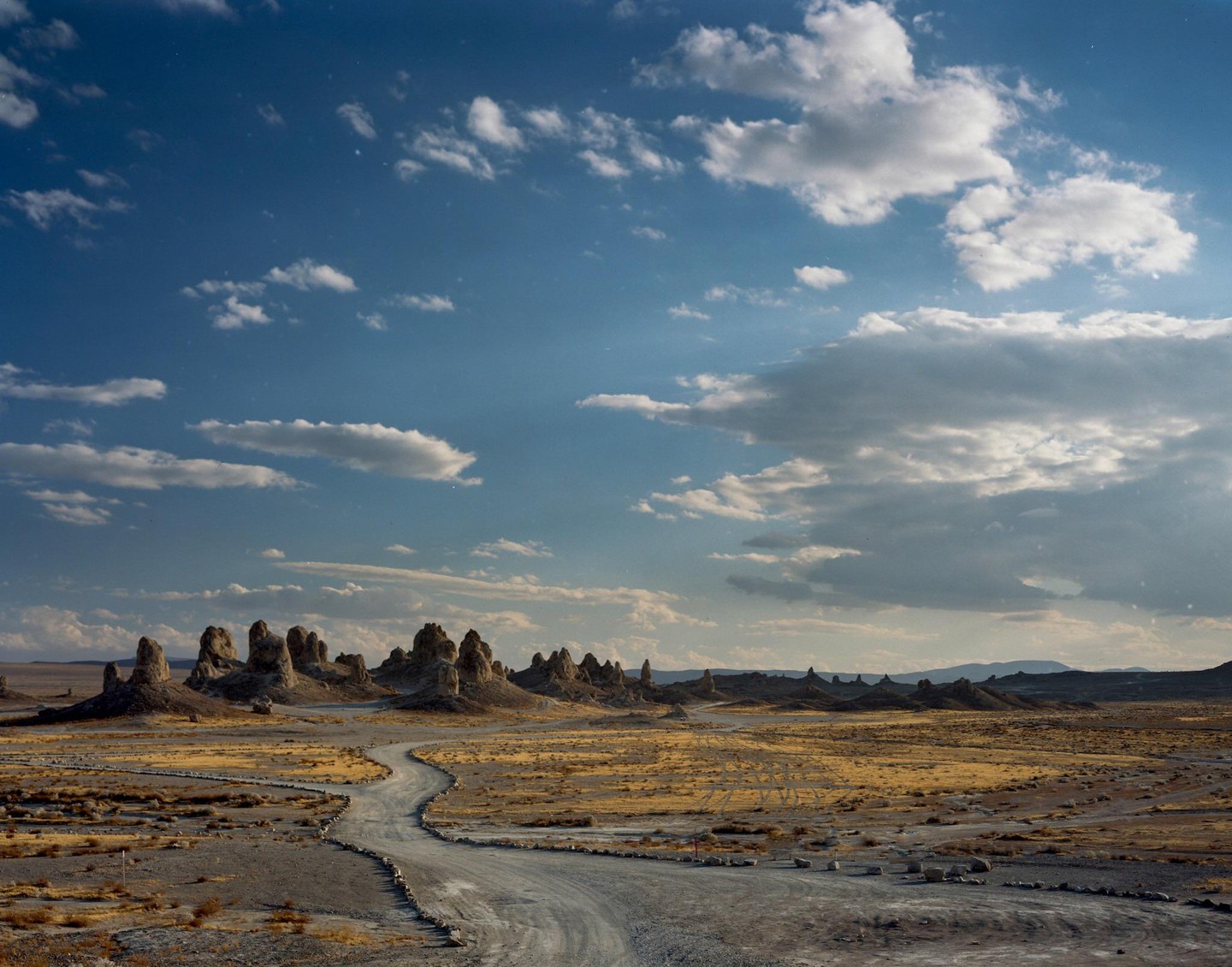 A winding dirt road leads through a desert landscape with rock formations under a partly cloudy sky.
