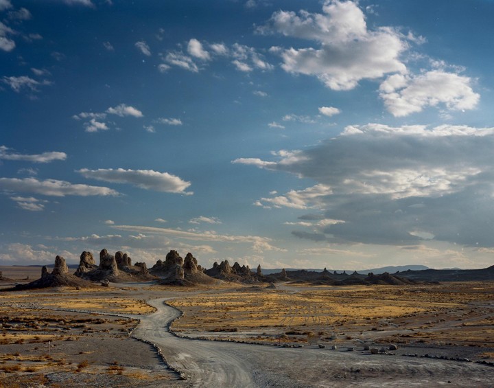 A winding dirt road leads through a desert landscape with rock formations under a partly cloudy sky.