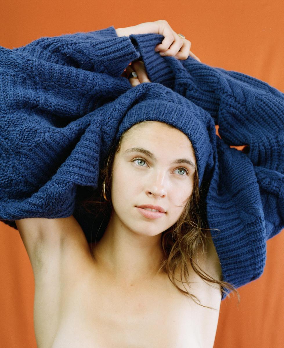 A woman with blue eyes pulls a blue knit sweater over her head against an orange background.