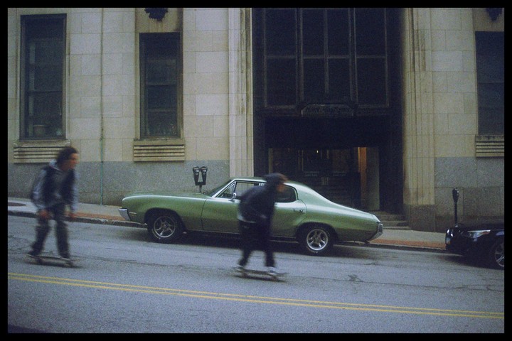 Two people on skateboards pass a green car parked on a city street.