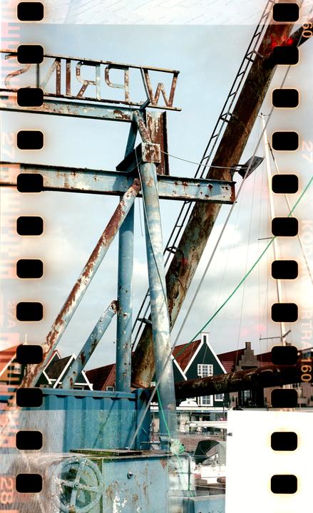 A rusty blue metal structure with a sign is visible against a cloudy sky, with traditional houses and a boat in the background.