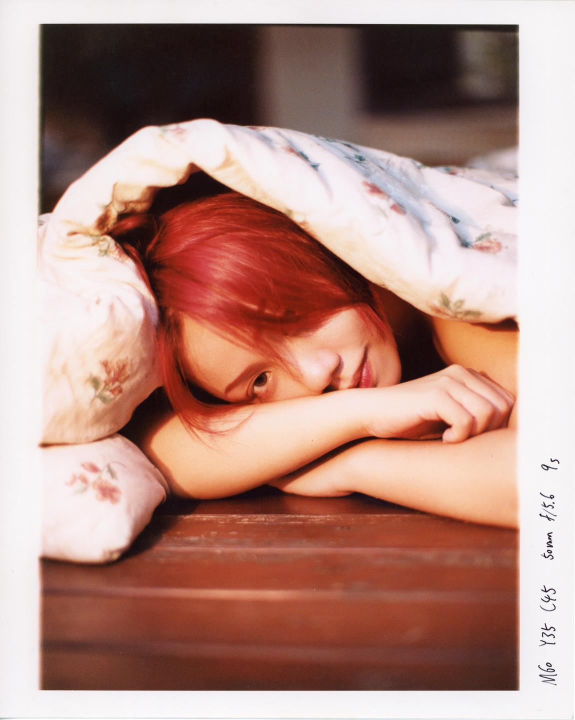 A woman with red hair peeks out from under a floral blanket, resting her head on her arms.