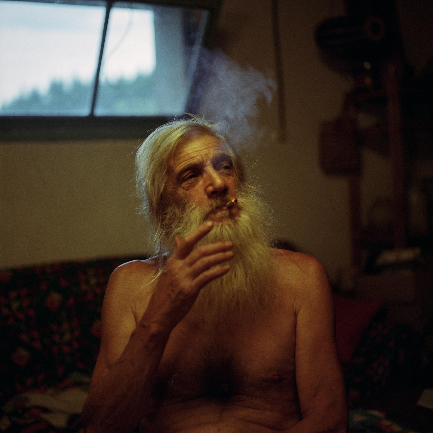 A man with a long white beard smokes a cigarette indoors.