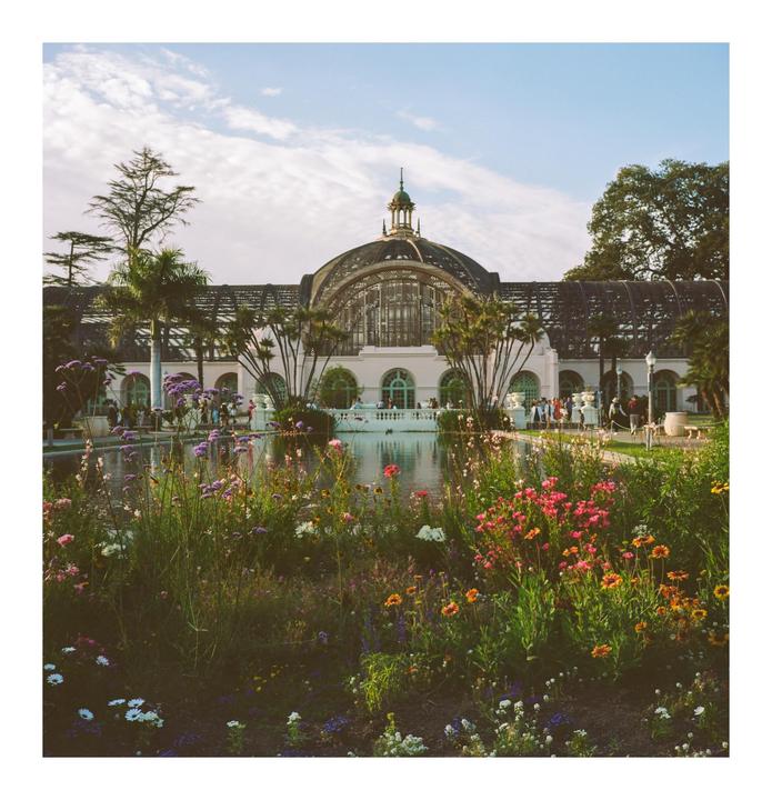A large domed building with a pond and colorful flowers in the foreground is visible under a partly cloudy sky.