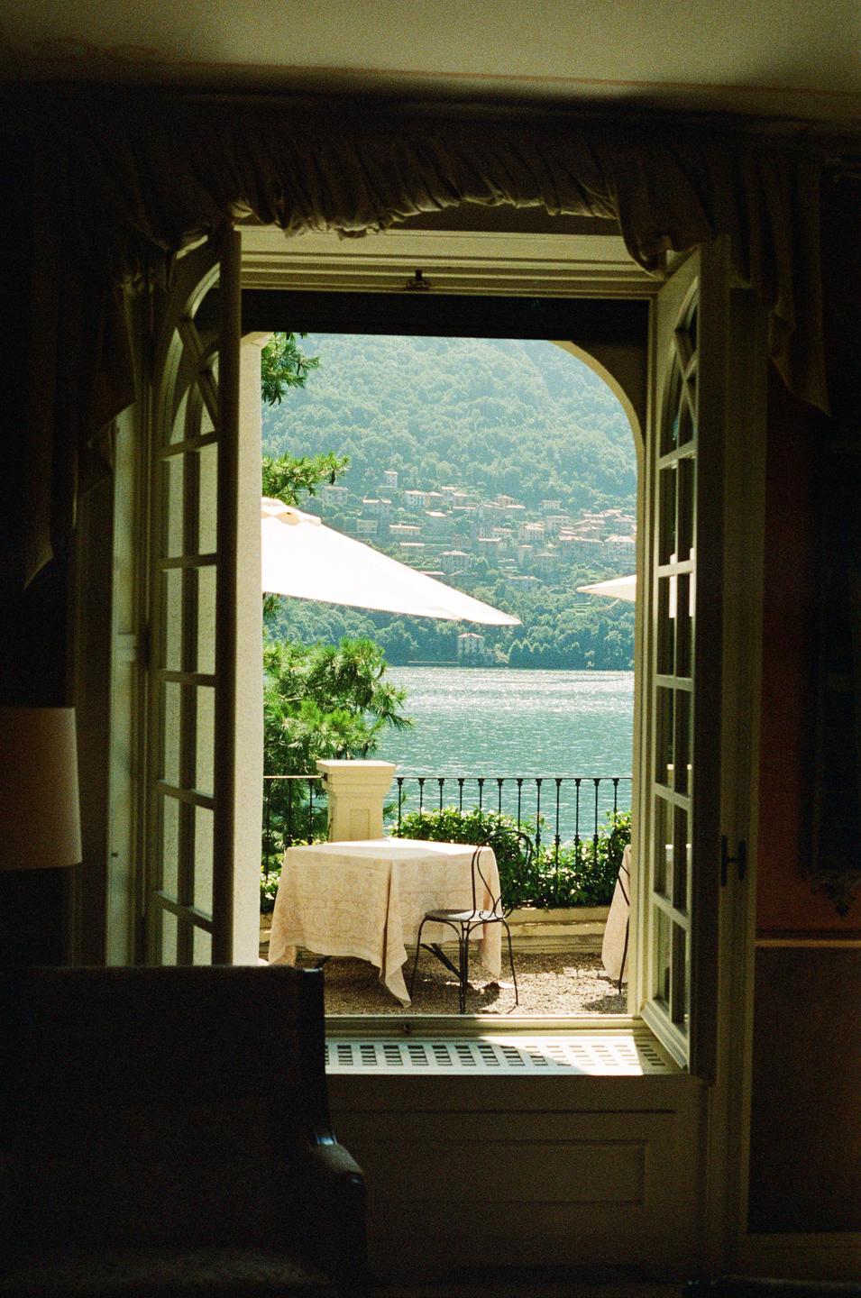 A view from an open door shows a patio with a table and chair overlooking a lake and a distant town on a hillside.