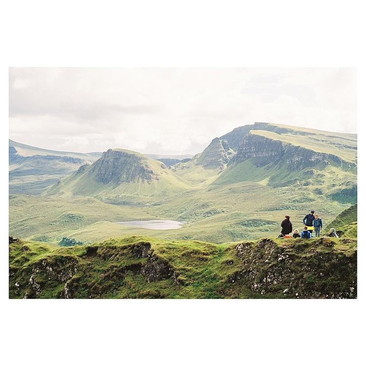 A group of people are gathered on a grassy hill overlooking a vast, mountainous landscape with a small lake.