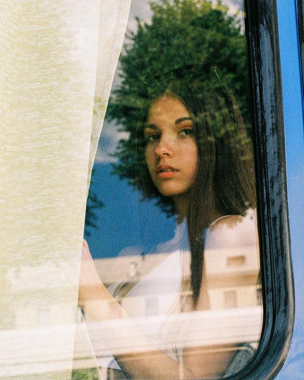 A young woman looks out a window with a tree and building reflected in the glass.