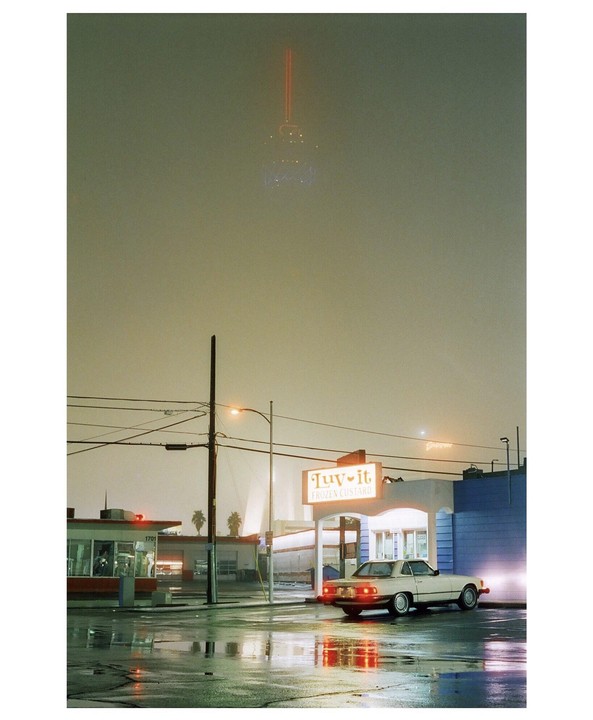 A white car is parked in front of a "Tuv-it Frozen Custard" shop on a wet street at night, with a distant tower visible in the hazy sky.