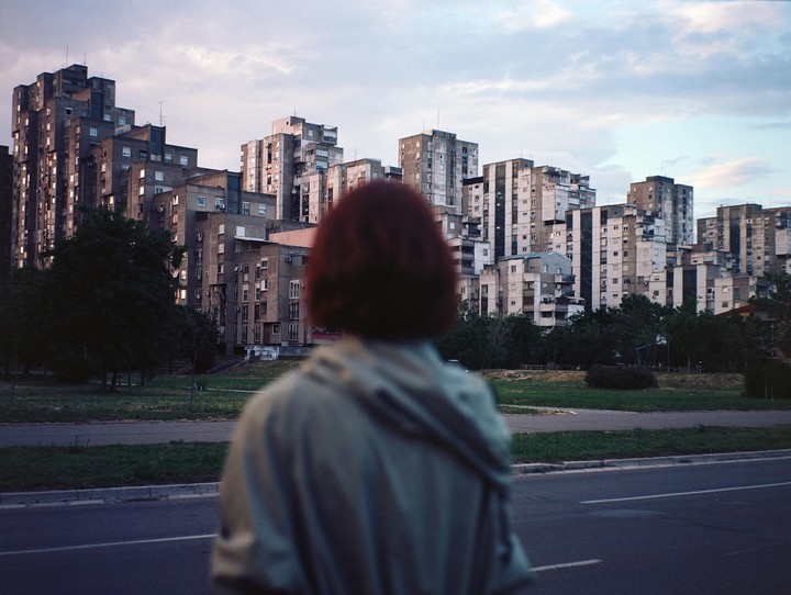 A person with red hair stands with their back to the viewer, looking at a city with many apartment buildings.
