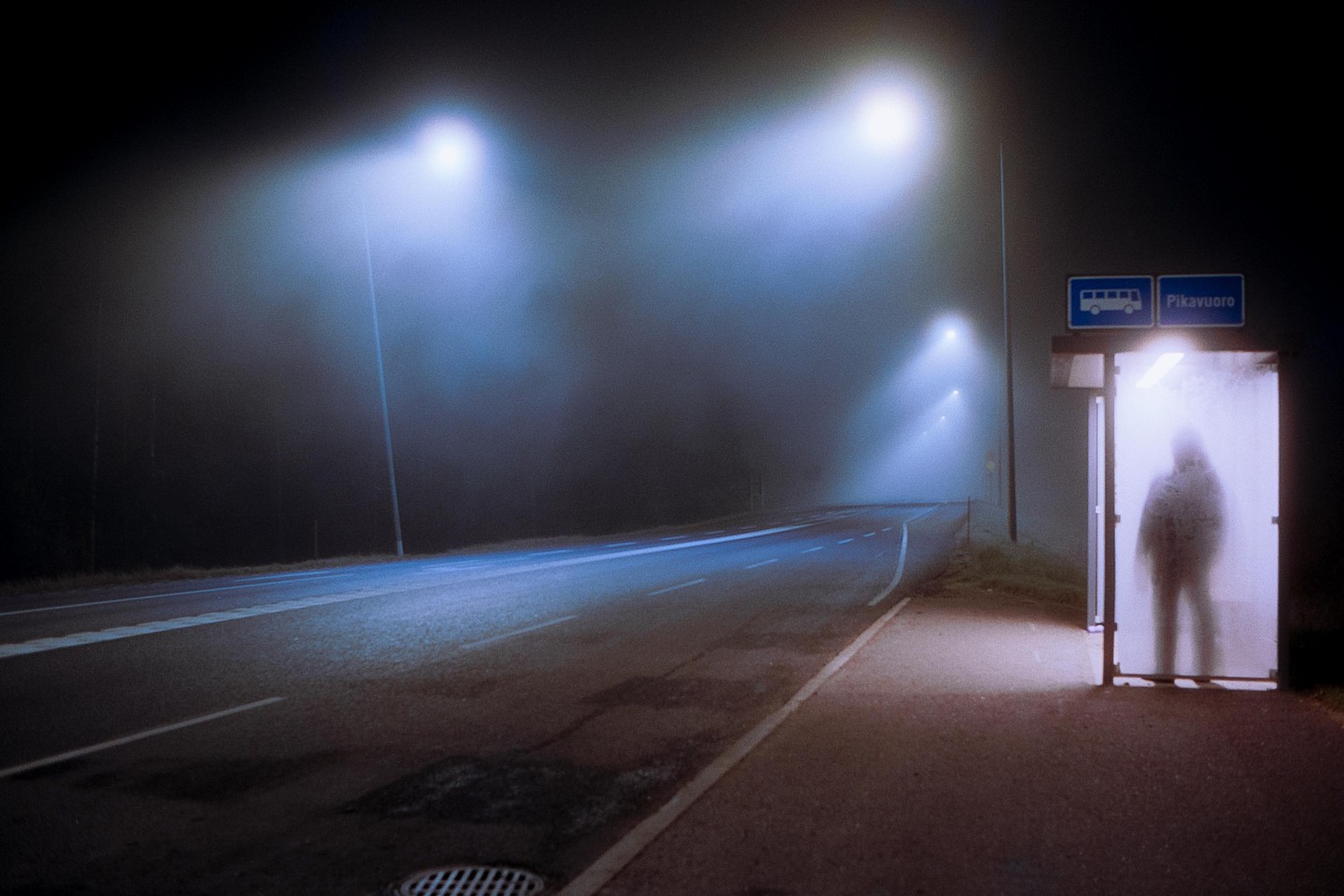 A person stands inside a bus stop shelter on a foggy night next to a road with streetlights.