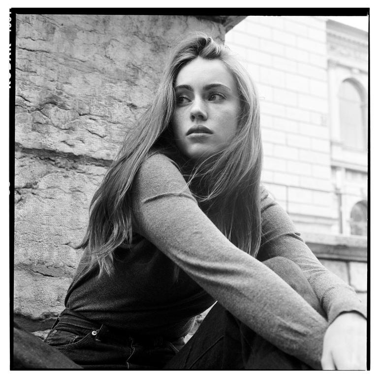 A young woman with long hair sits outdoors next to a stone wall, looking away from the camera.