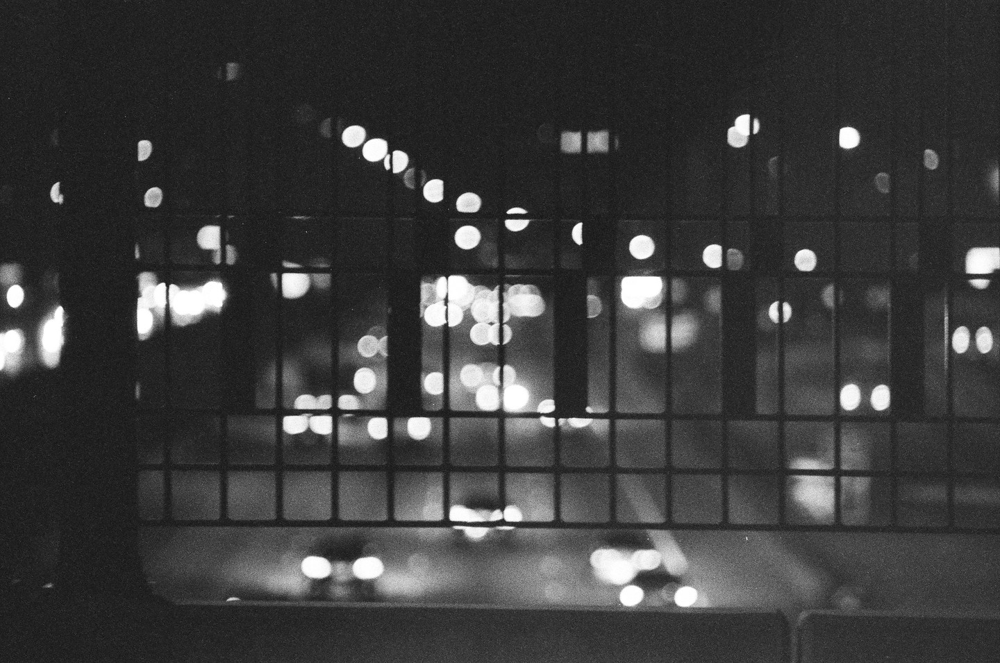 A black and white photo shows a blurry street at night through a metal fence.
