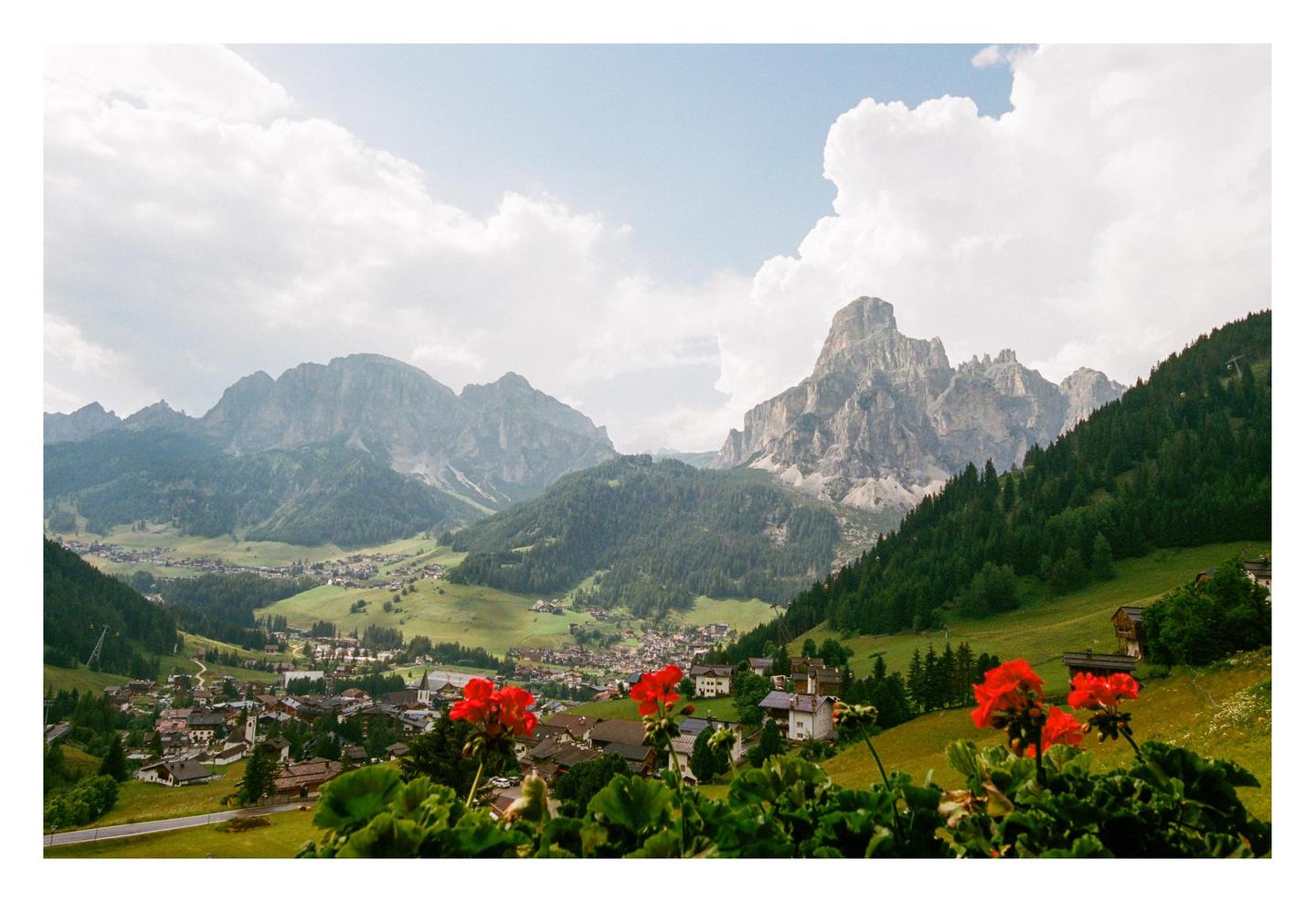 Red geraniums frame a view of a mountain village nestled in a valley with towering peaks under a cloudy sky.