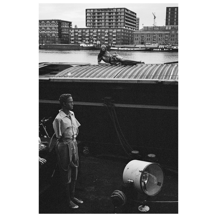 A man stands on a boat deck while a mannequin lies on the boat roof with buildings and water in the background.