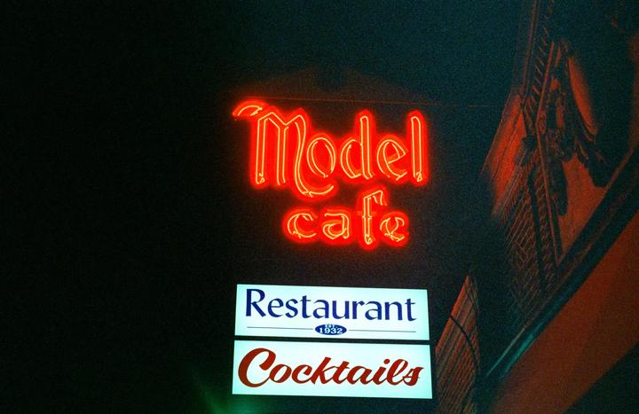A neon sign for "Model Cafe" is visible above signs for "Restaurant" and "Cocktails" at night.