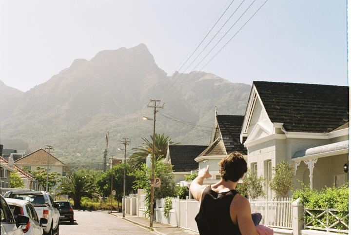 A man in a black tank top points towards a hazy mountain in the distance, with houses and parked cars lining a street in the foreground.