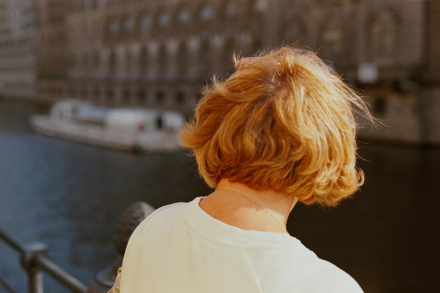 A woman with short, curly, reddish-blonde hair stands with her back to the viewer, looking out over a body of water with a boat and buildings in the background.