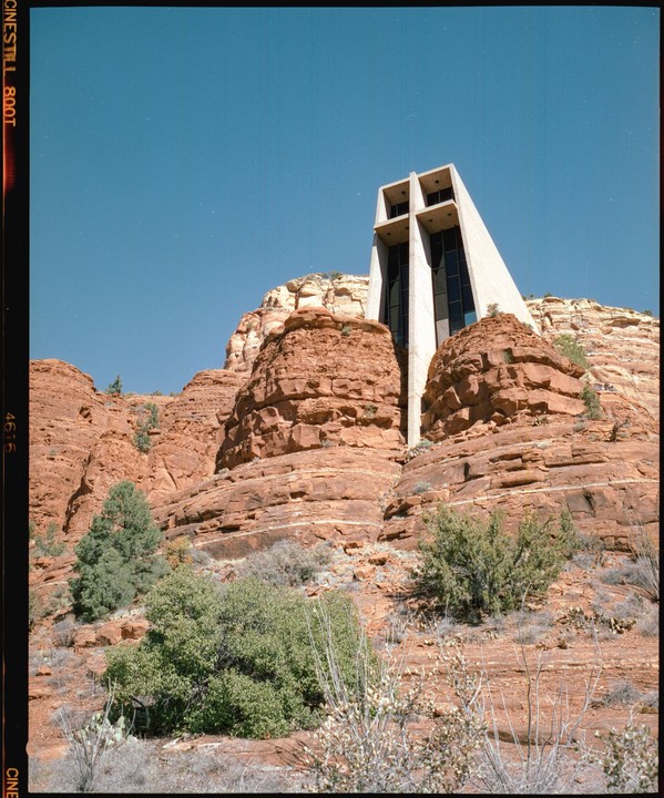 A modern concrete church with a cross-shaped facade is built into a red rock formation under a clear blue sky.