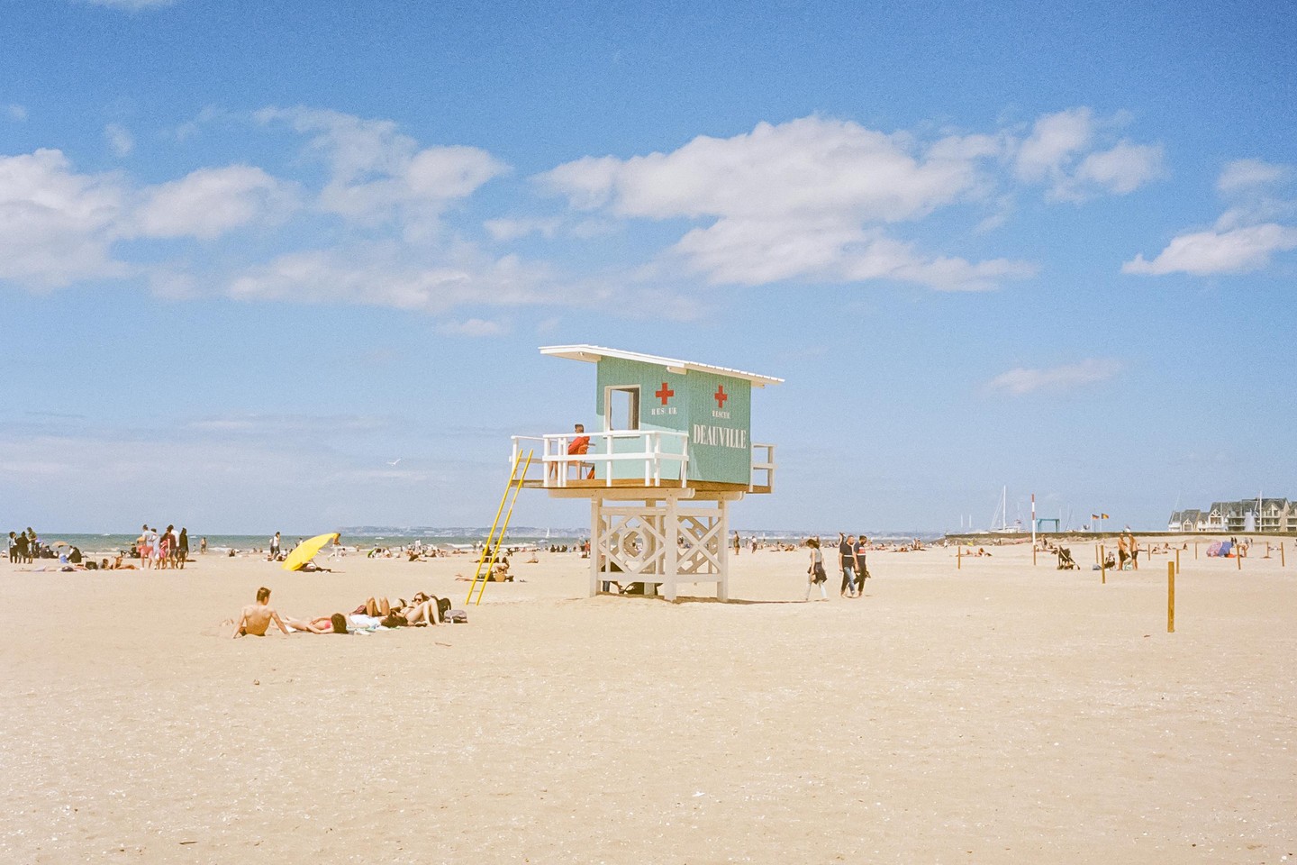 A lifeguard tower stands on a crowded sandy beach under a blue sky with scattered clouds.