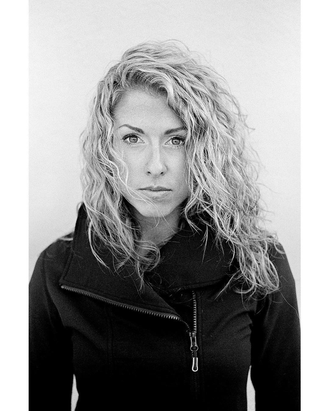 A woman with curly hair and a black jacket looks directly at the viewer.