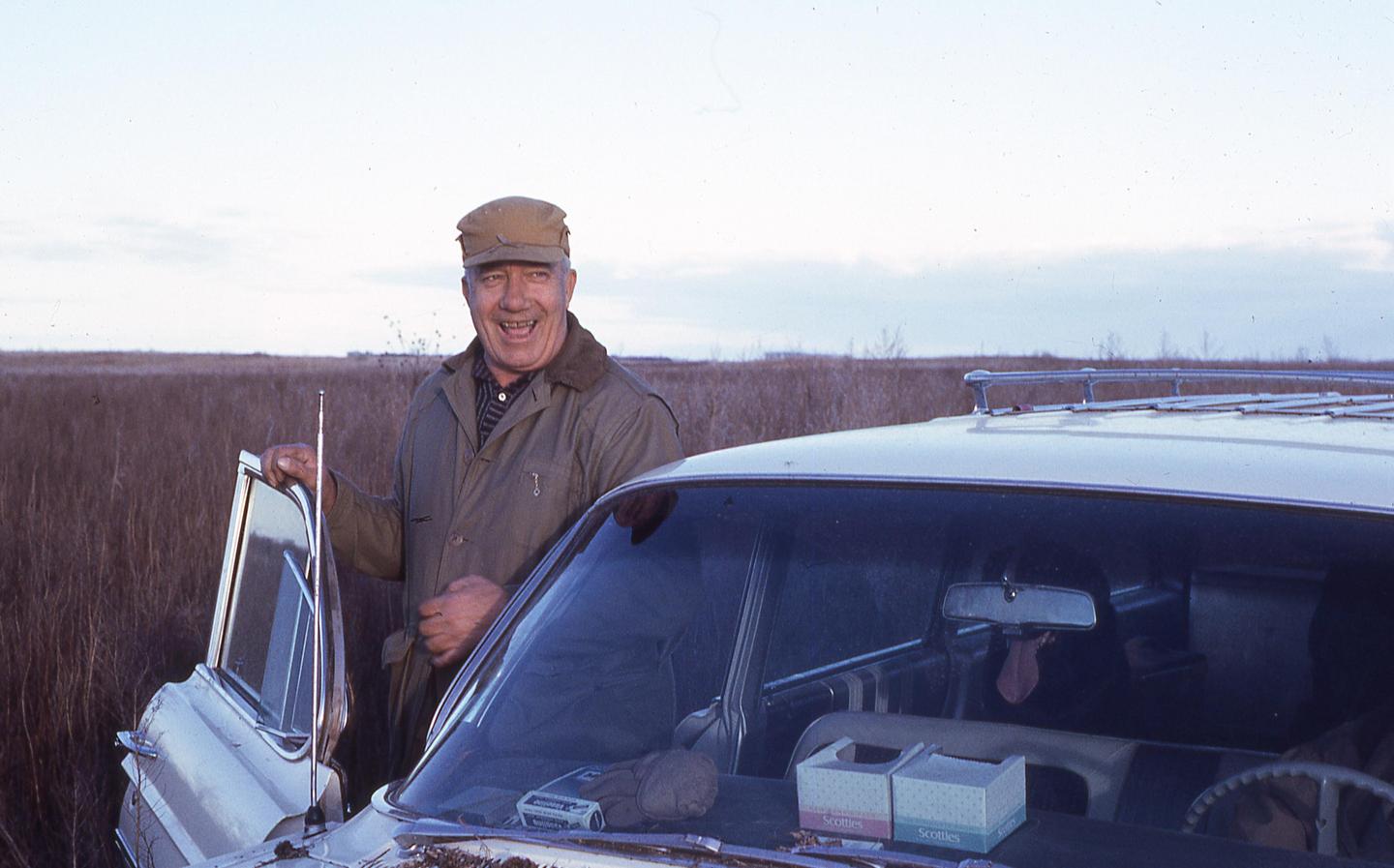 A man in a cap and jacket stands next to a white car with an open door, in a field of tall dry grass.