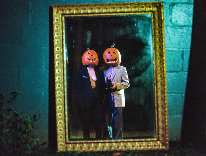 Two people wearing pumpkin masks are reflected in a large, ornate mirror placed outdoors at night.