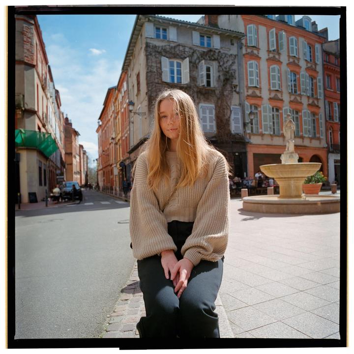 A woman sits on a curb in a European city with a fountain and buildings in the background.