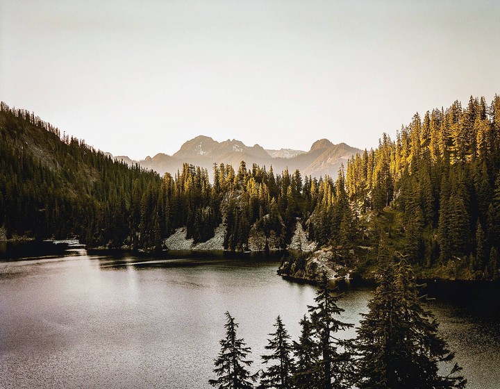 A lake is surrounded by a dense forest with mountains in the background under a bright sky.