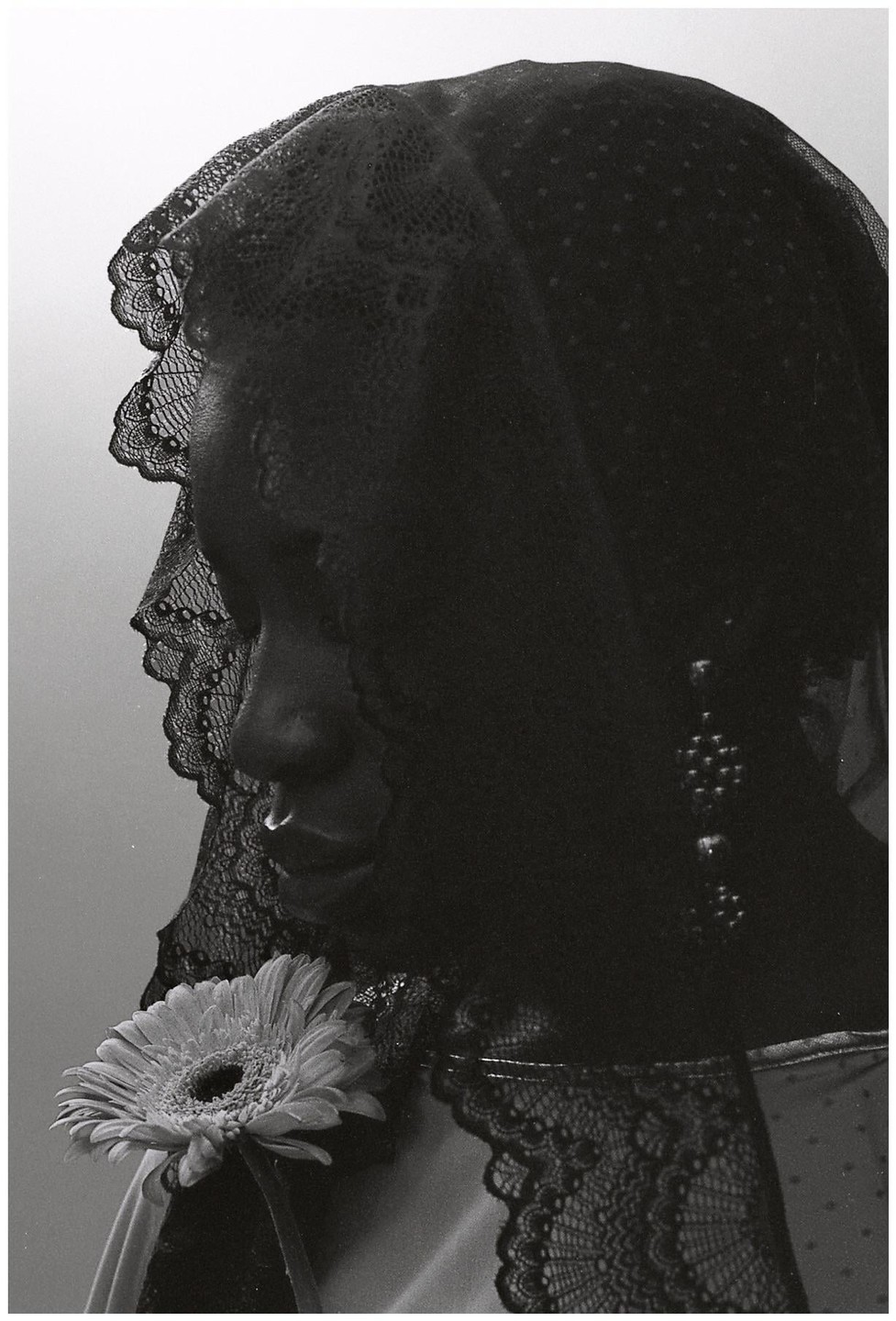 A woman with a nose ring and a lace veil holds a gerbera daisy.