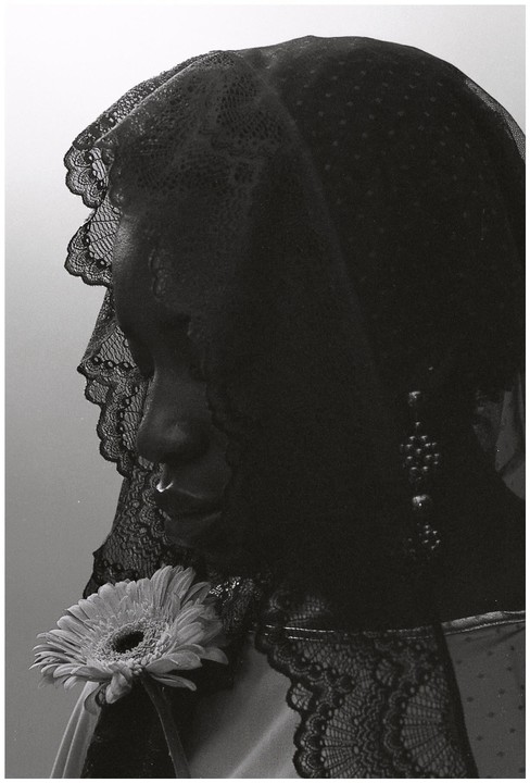 A woman with a nose ring and a lace veil holds a gerbera daisy.