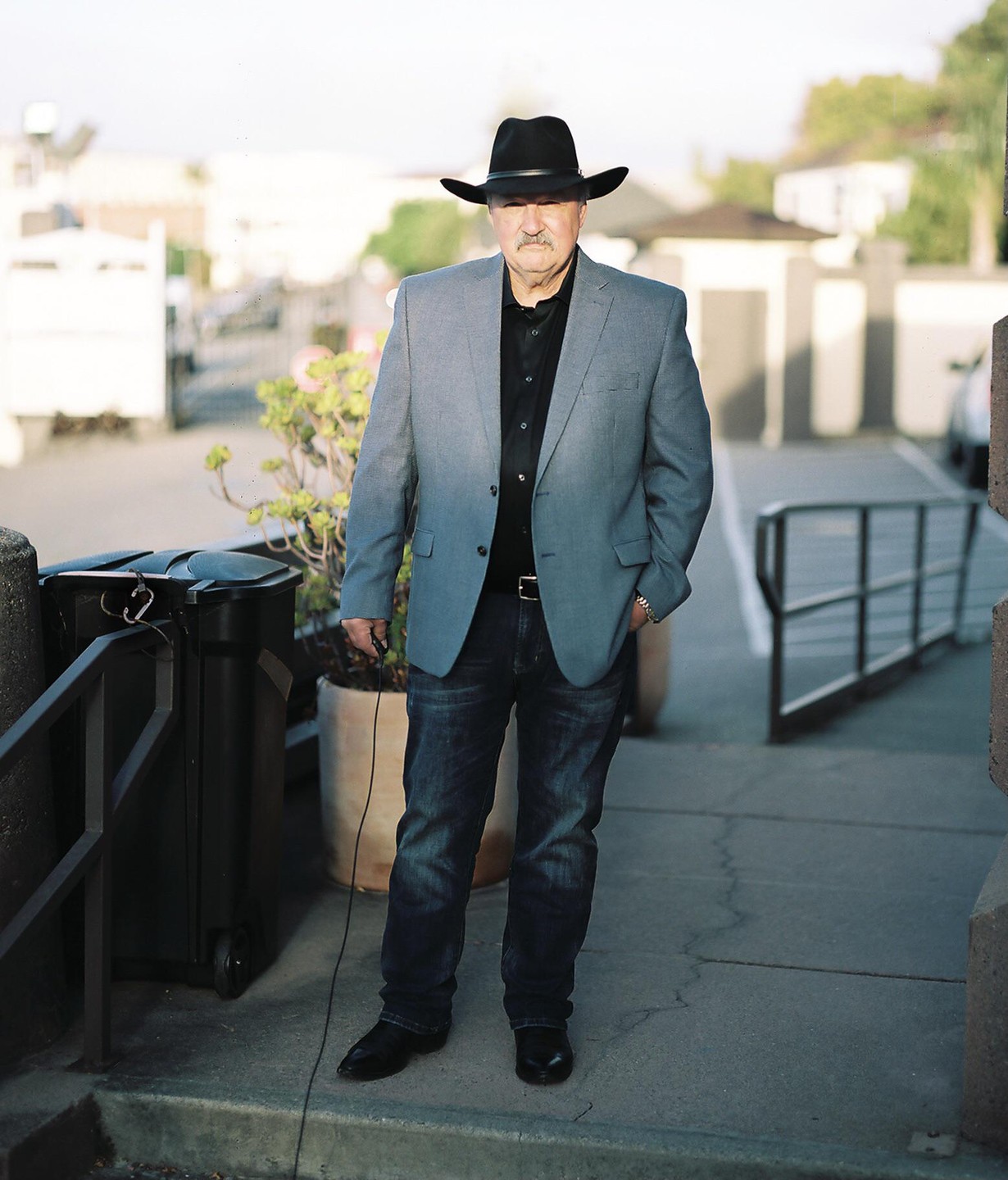 A man wearing a cowboy hat, blazer, and jeans stands on a sidewalk.