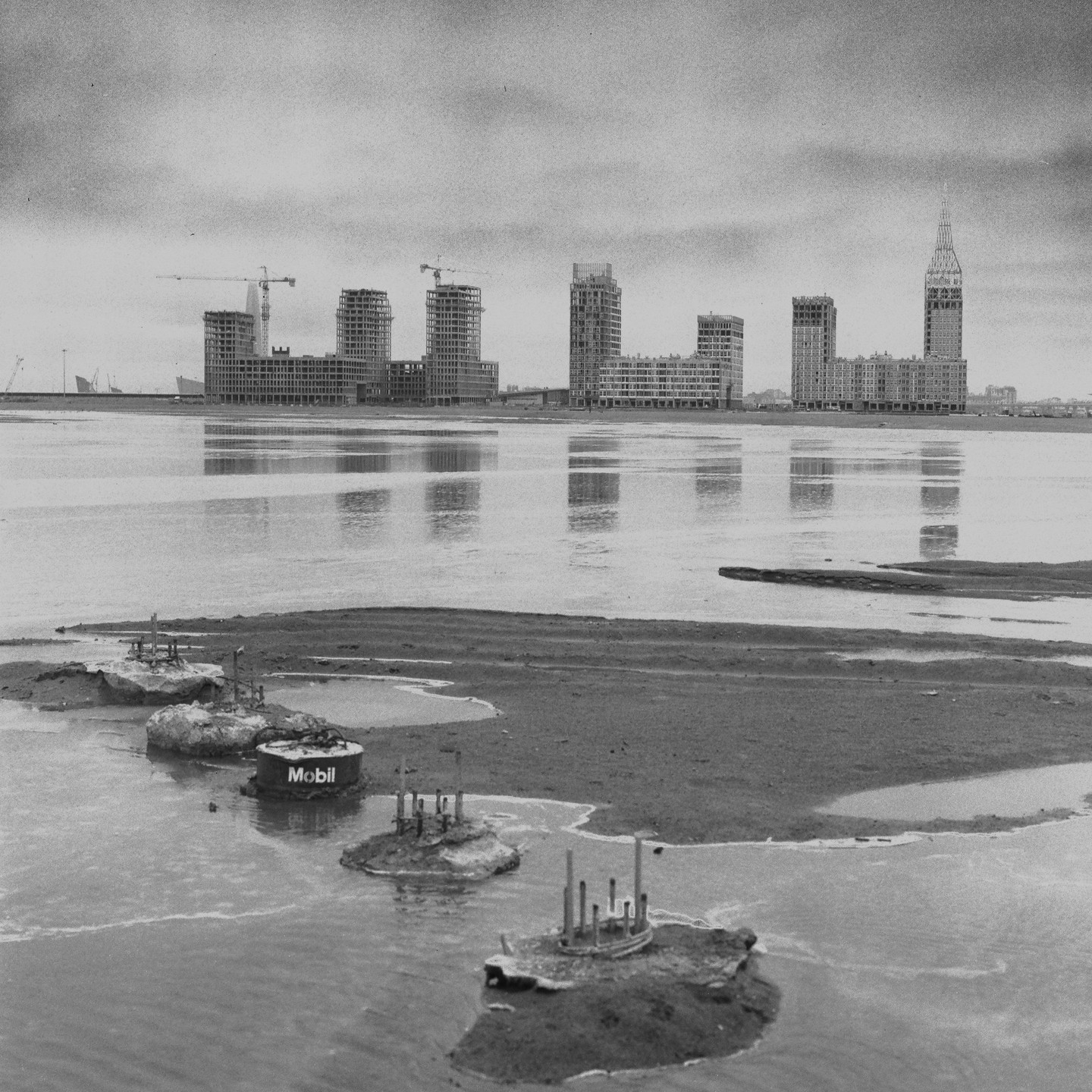 A black and white landscape shows a row of buildings under construction across a body of water, with a Mobil barrel in the foreground.