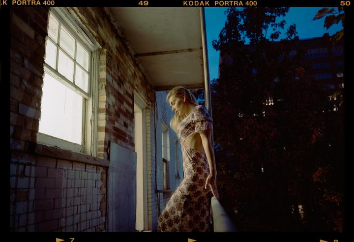 A woman in a floral dress stands on a balcony next to a brick wall and window during blue hour.