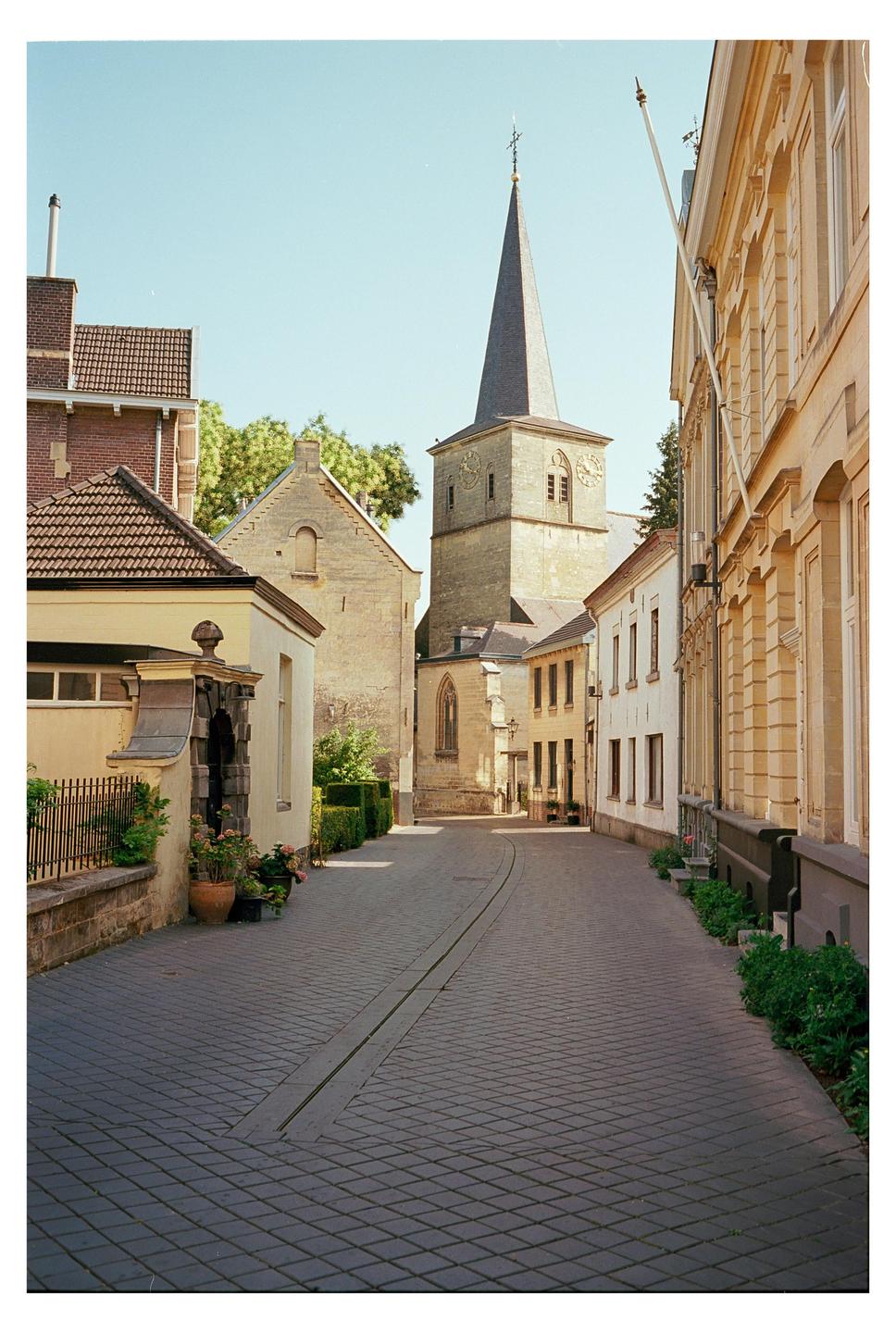 A narrow cobblestone street curves through a historic town with a tall church steeple visible in the background.