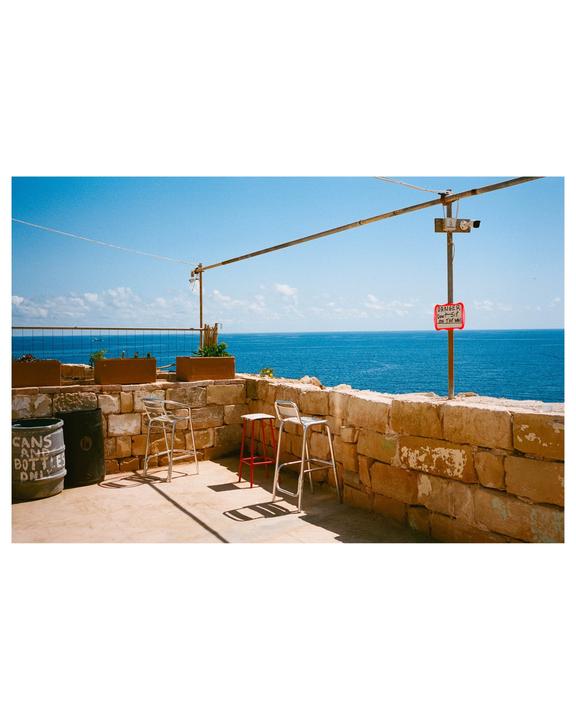 Three bar stools sit on a stone patio overlooking a bright blue sea under a clear sky.