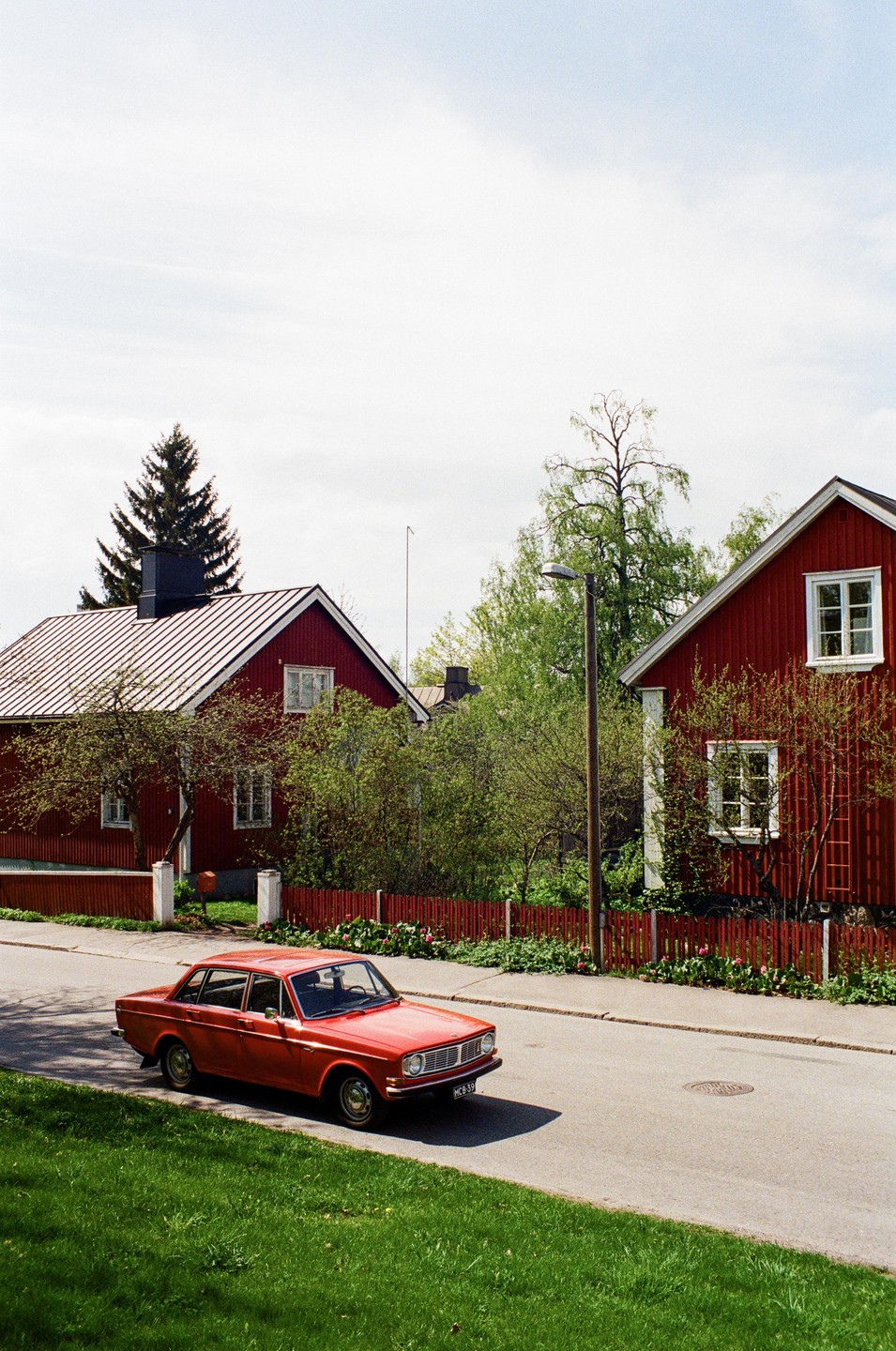 A red vintage car is parked on the side of a street lined with red houses and green trees under a bright sky.
