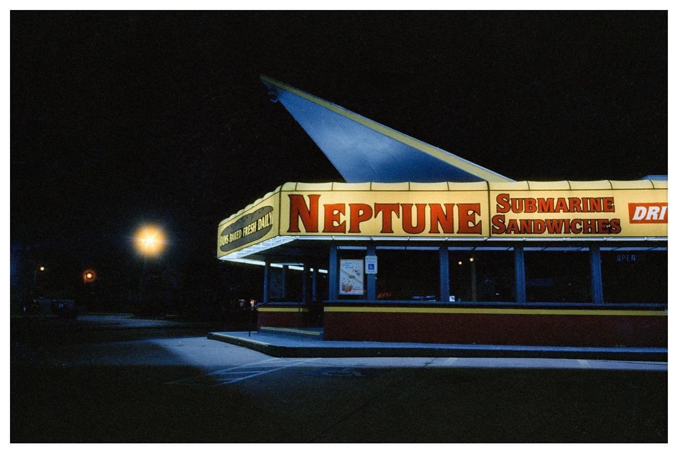 A brightly lit Neptune Submarine Sandwiches restaurant stands out against a dark night sky.