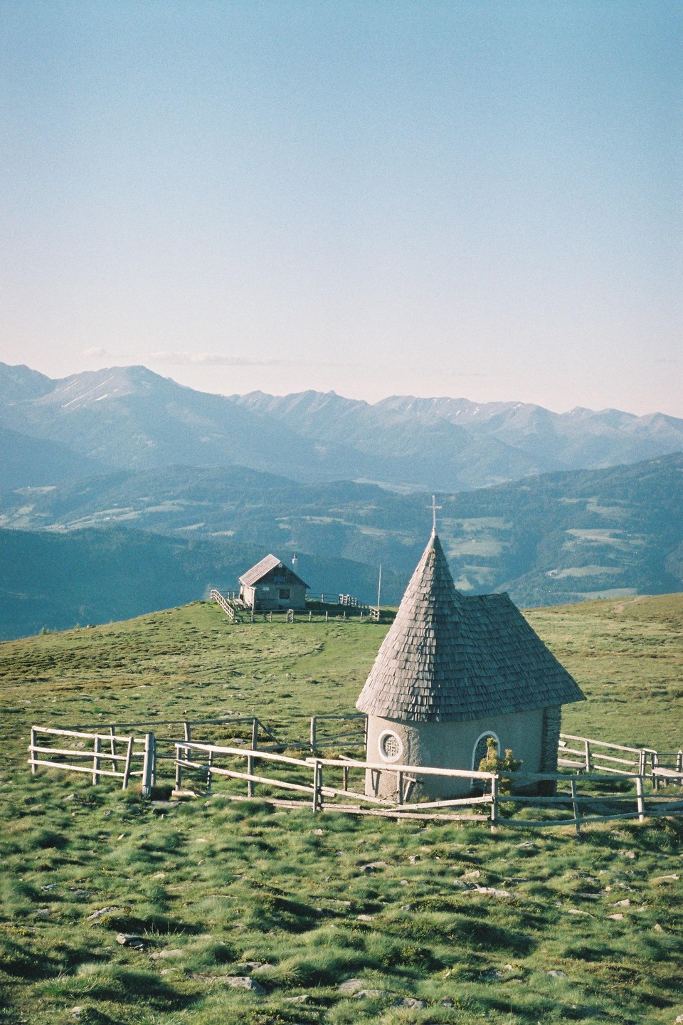 A small chapel with a conical roof and a wooden fence stands on a grassy hill with mountains in the background.
