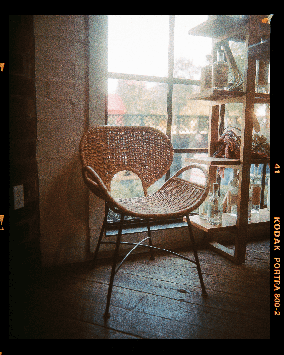 A wicker chair sits on a wooden floor next to a window and a wooden shelf with bottles.