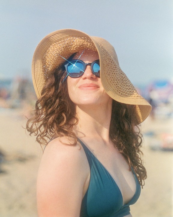 A woman wearing a straw hat and sunglasses smiles while standing on a beach.